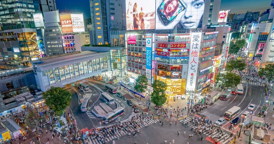 夕暮れの渋谷スクランブル交差点を上から見た様子。光るビジョンに覆われたビルの間を人波が渡っています