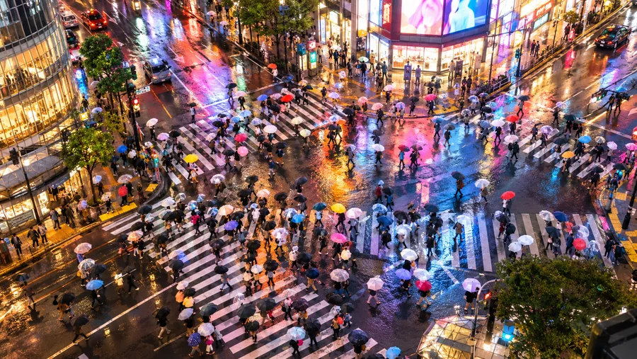 Bunte Regenschirme auf einer riesigen Diagonalkreuzung in Tokio im Regen, Neonlicht spiegelt sich auf der nassen Straße